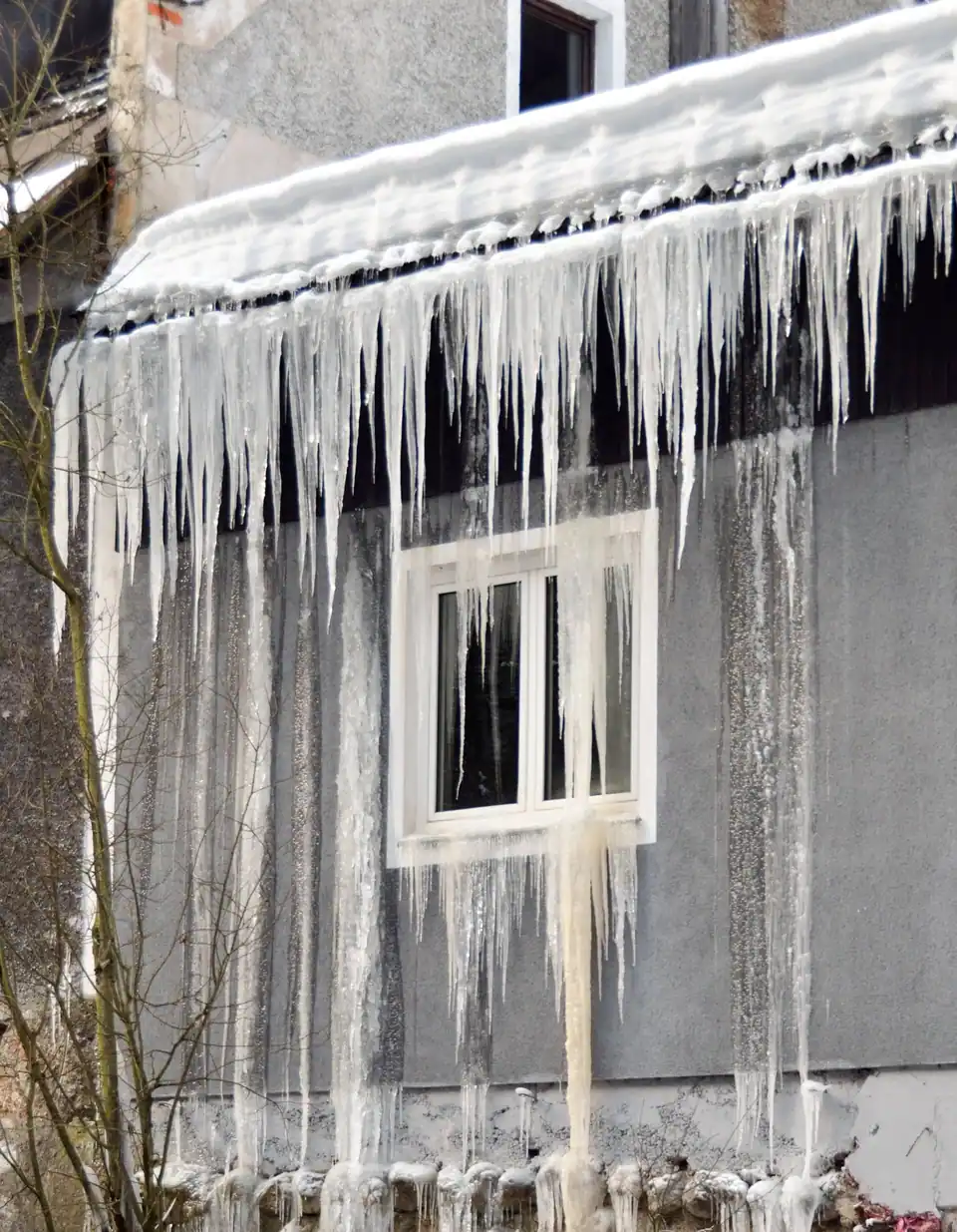 Thick icicles and frozen meltwater hanging from a snowy roof edge down a house wall past a window