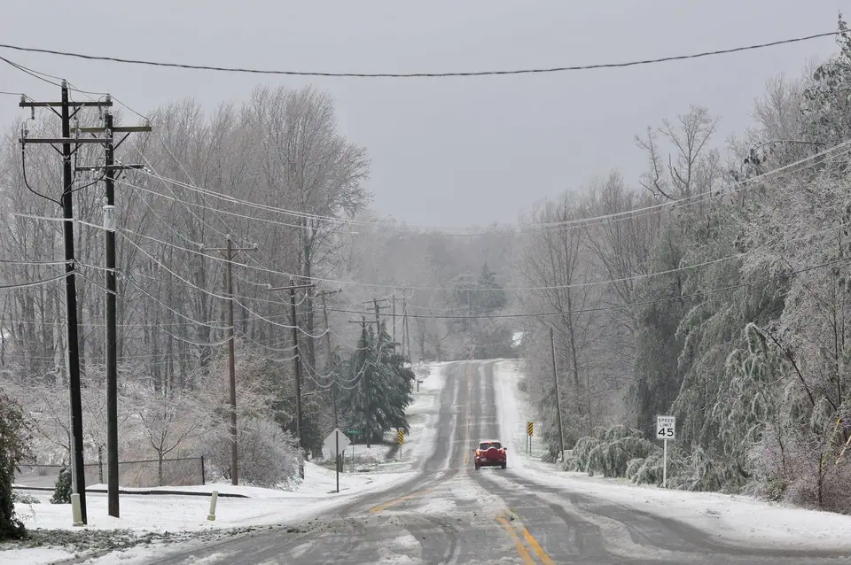 An ice-coated country road lined with power lines and bent trees during a winter storm