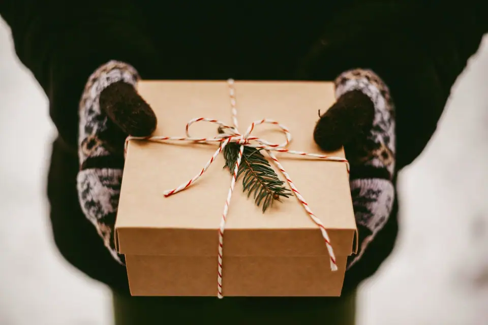 Hands in knitted mittens holding a kraft paper gift box tied with twine and a pine sprig