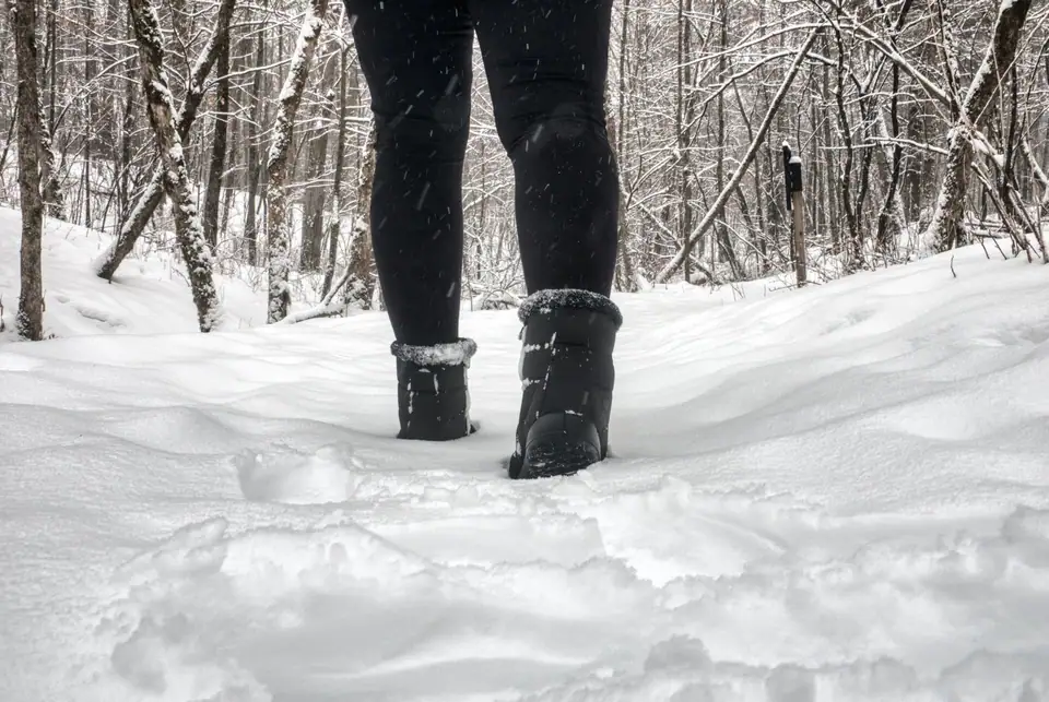A person in dark pants and snow boots walking along a snowy forest trail, seen from behind below the knee