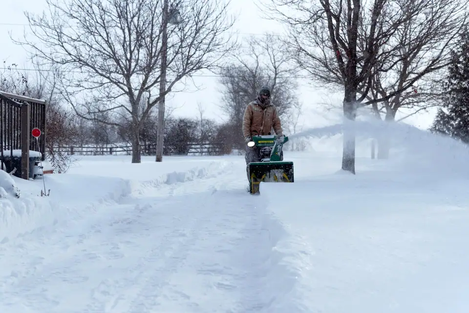 A snow blower clearing a snow-covered driveway lined with bare trees