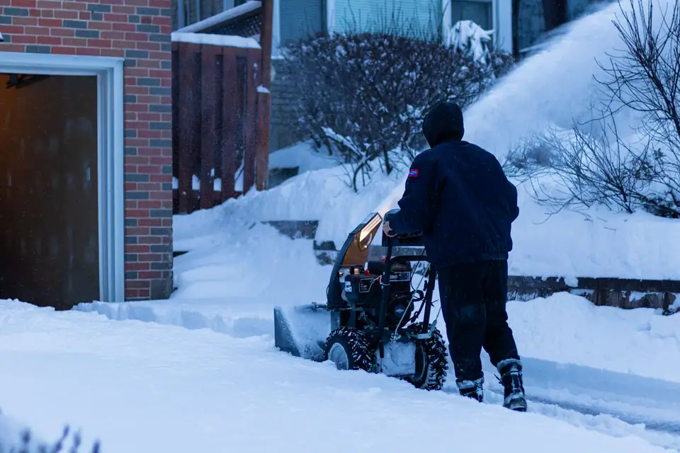 A person pushing a snowblower along a snowy sidewalk at dusk
